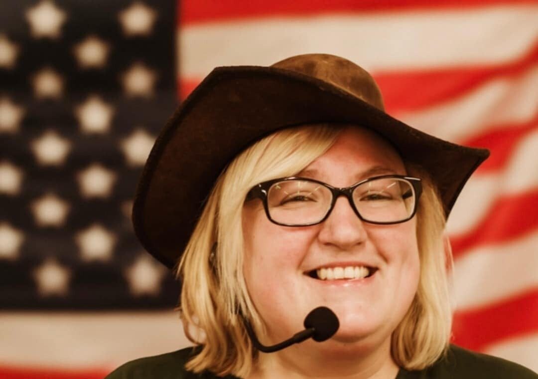 Carina, the line dancing instructor, smiling in a brown cowboy hat and headset microphone in front of an American flag