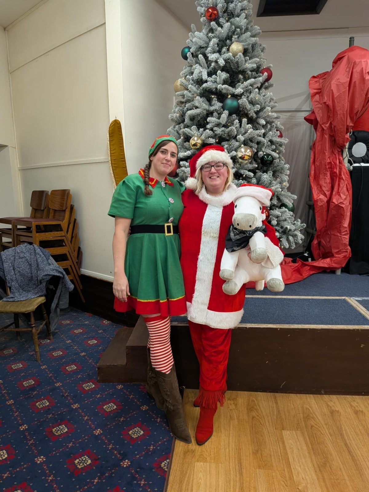Carina dressed as Santa holding the group's plush white horse mascot, beside a helper in an elf costume in front of a Christmas tree