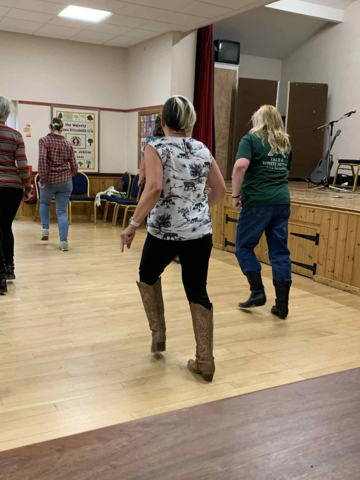 Dancers in cowboy boots mid-routine on a wooden hall floor, one wearing a green Vale and White Horse Line Dancers t-shirt
