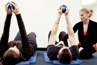 A class lying on mats raising small exercise balls overhead while Caroline instructs
