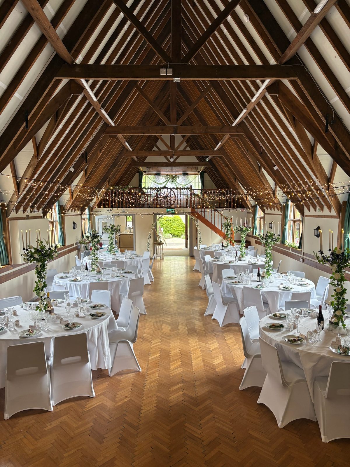 Round wedding tables dressed in white, with greenery along the gallery rail and the hall doors open to the garden
